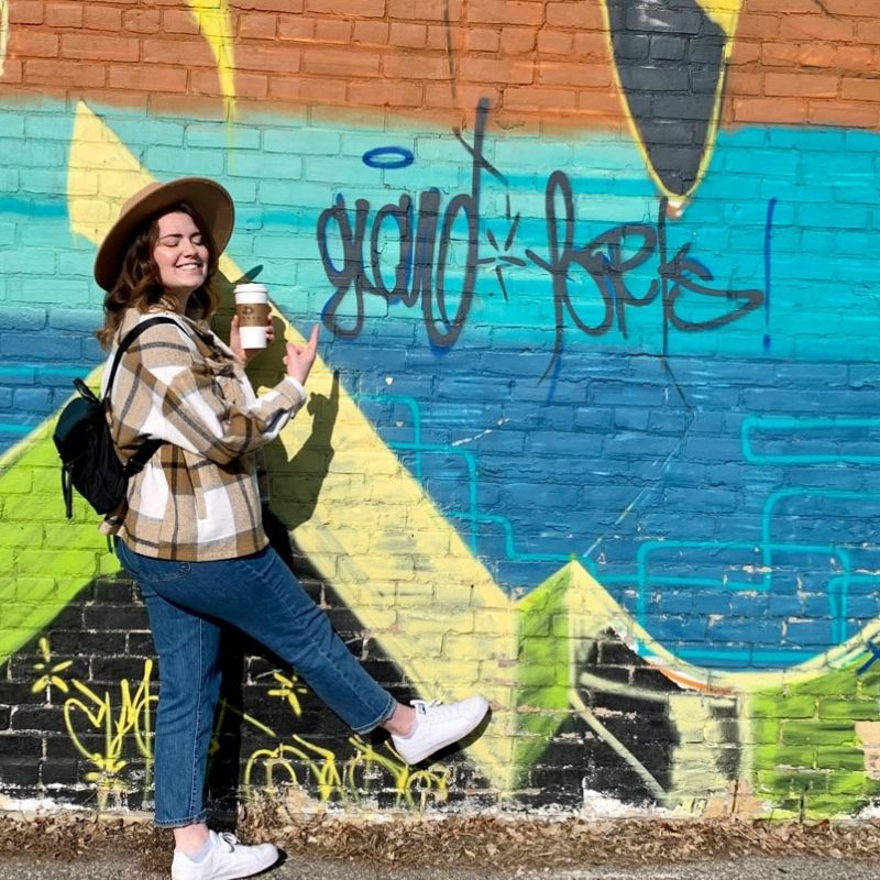 Emily exploring Grand Forks in front of a graffiti wall pointing to the words 