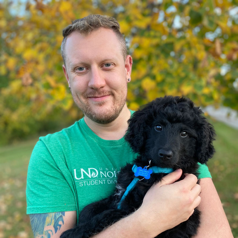 Jeff wearing a UND shirt, holding his black long haired puppy on the Greenway in Grand Forks, ND