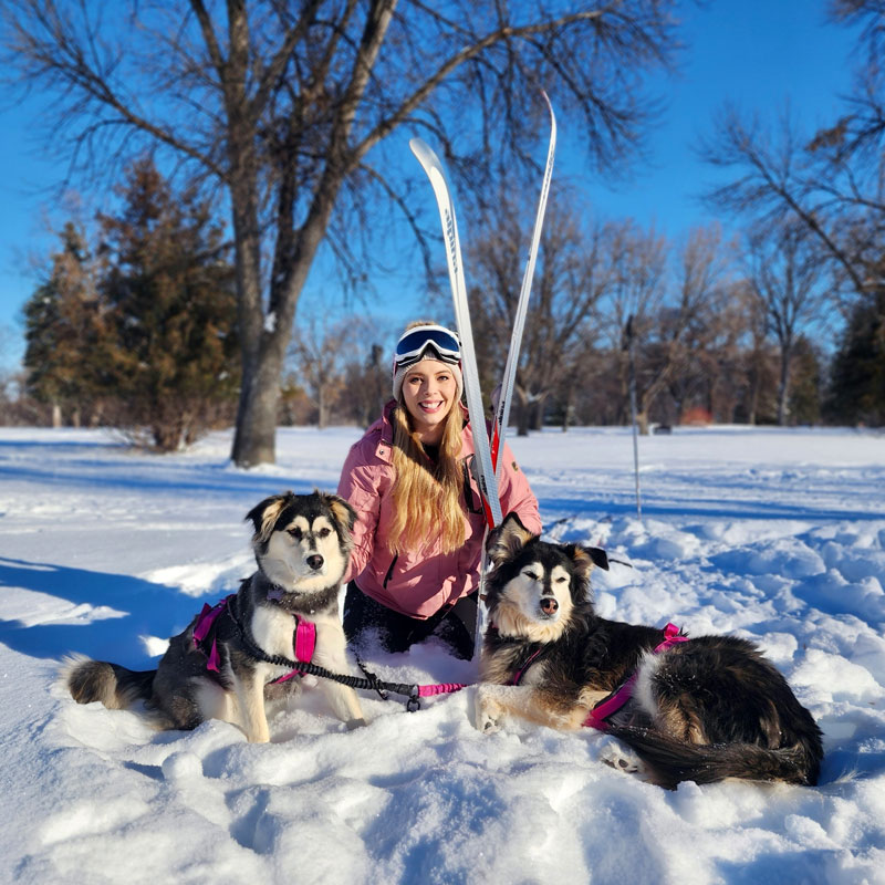 Woman poses with skis and two dogs in snow in Grand Forks, ND.