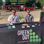 Two men sit behind table covered by table cloth that reads "Green Dot" in Grand Forks, ND