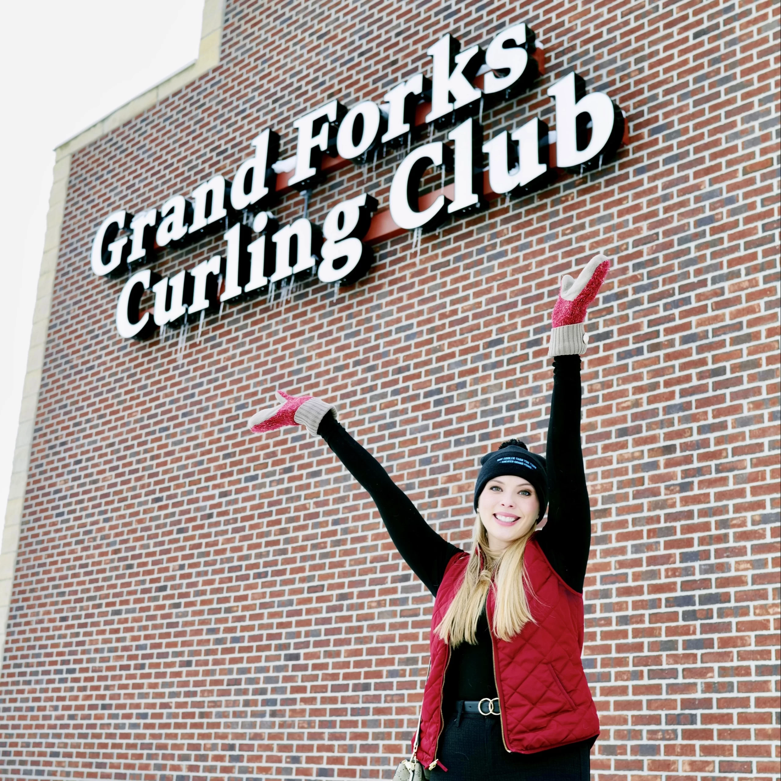 Winter in Grand Forks Cassandra standing in front of brick building that reads, 'Grand Forks curling club'