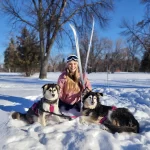 Winter in Grand Forks Cassandra sitting with her skiis and two dogs in the snow.