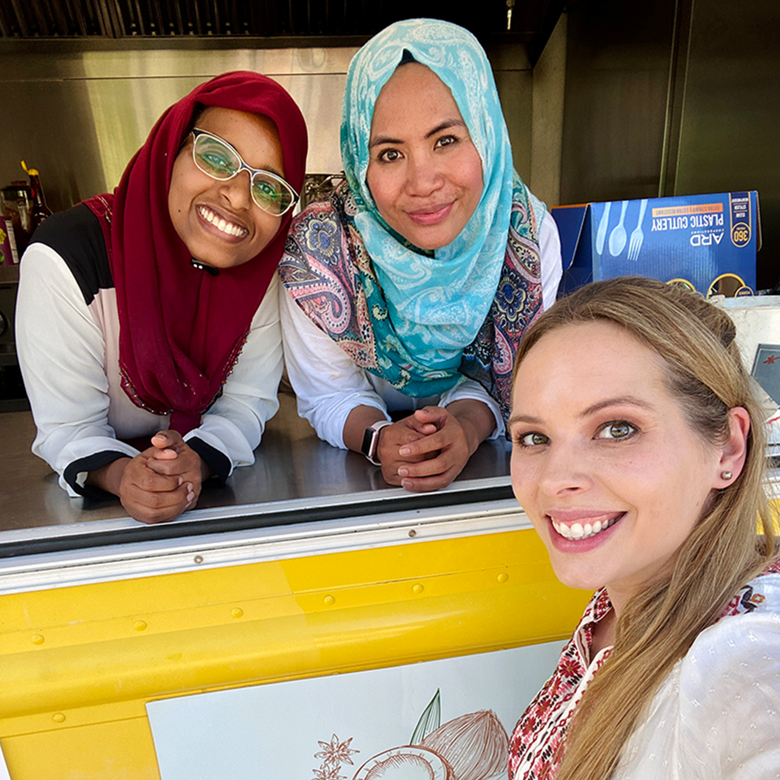 Three woman smiling for a photo, two working in a food truck, one on the outside taking the photo.