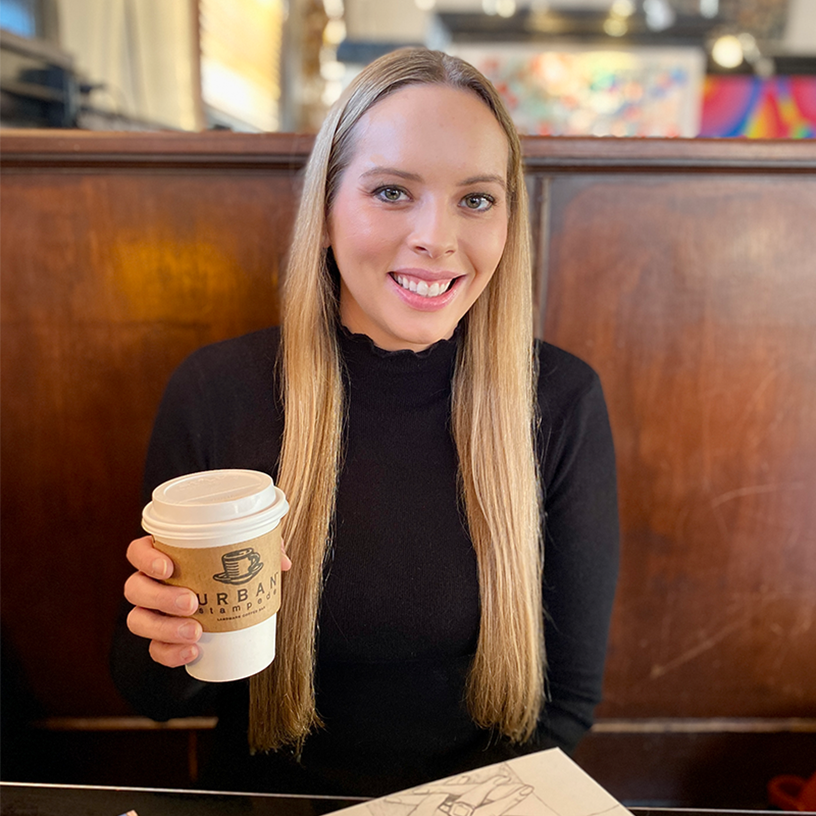 Woman in a booth at a coffee shop, holding a warm beverage and smiling for a photo.
