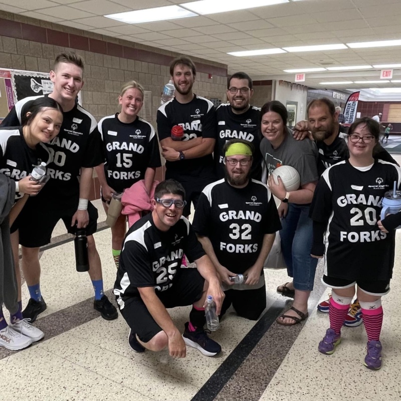 The Grand Forks Special Olympics volleyball team wears black uniforms and stand in a group in a hallway