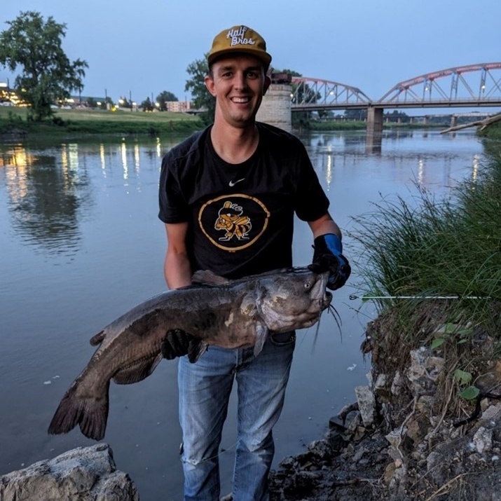 A man holds a large catfish with Sorlie Bridge in the background in Grand Forks