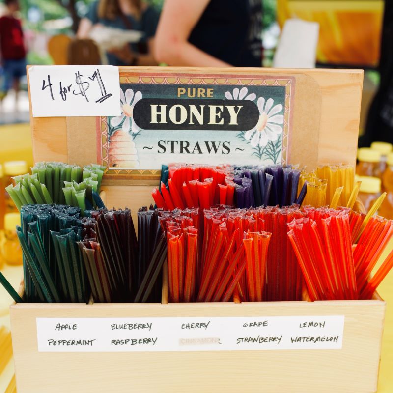 A wooden stand holds different colored sticks filled with flavored honey. A sign reads "Pure Honey Straws: 4 for $1"