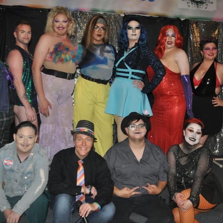 A group of individuals in drag stand in front of BJ Armani's Cabaret banner at a local pride show.