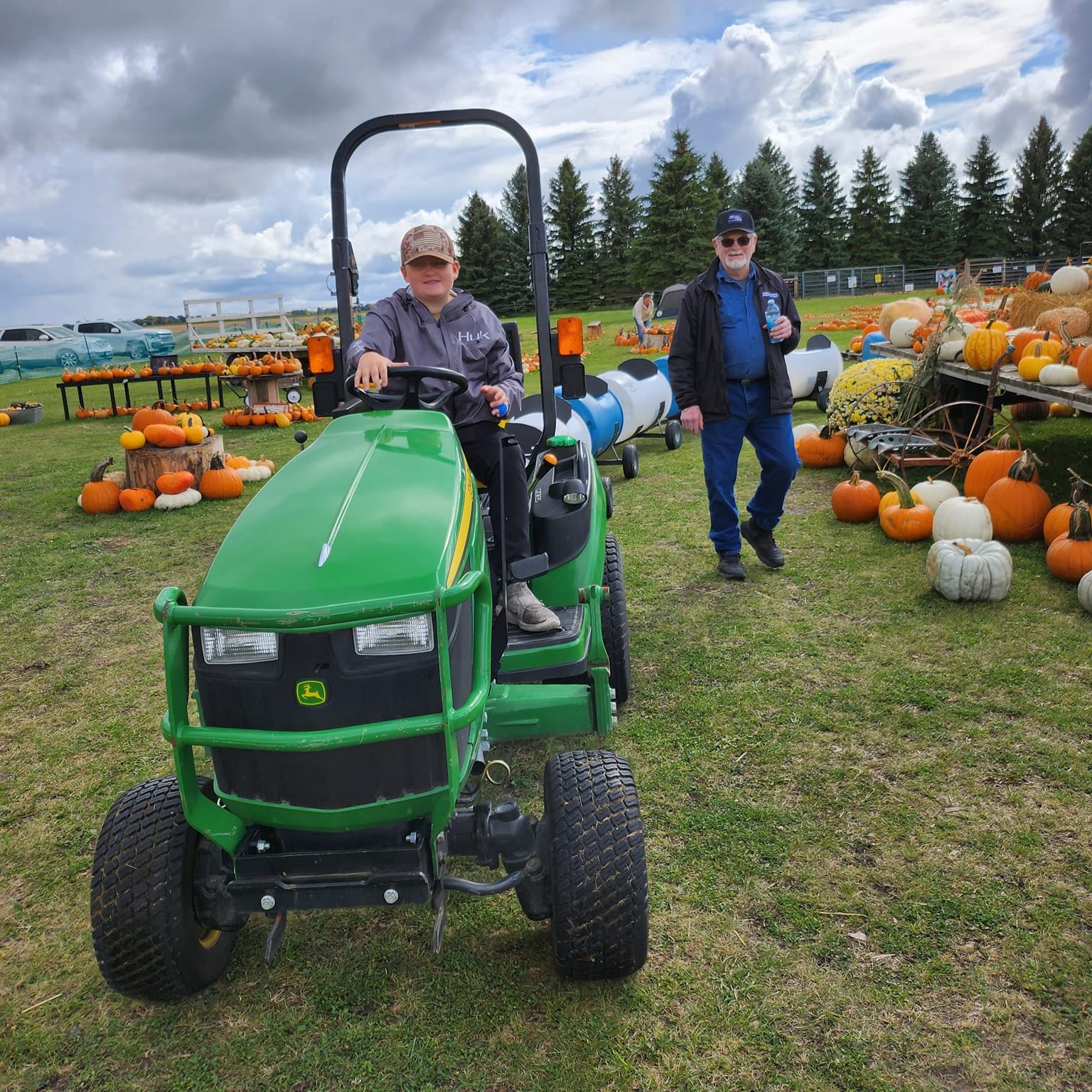 A young gentleman rides a green John Deer tractor that hauls a train of cars for children to sit in. He passes in front of a background of pumpkins while another gentleman looks on.