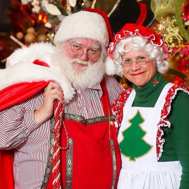 Santa and Mrs. Clause smile cheerfully in front of a festive scene.