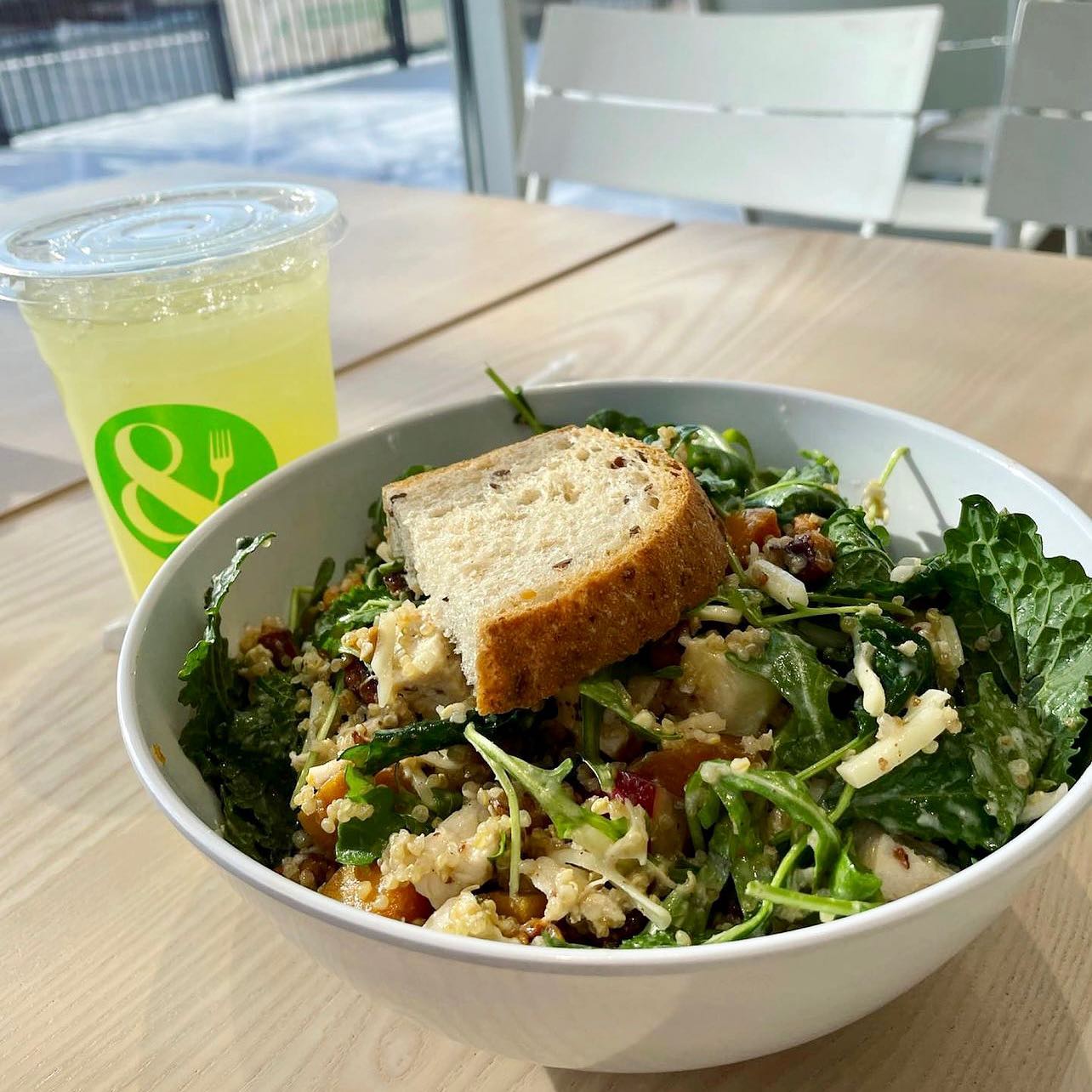 A salad with quinoa and a baguette slice on top sit on a brightly lit table next to a lemonade donning the Crisp and Green logo