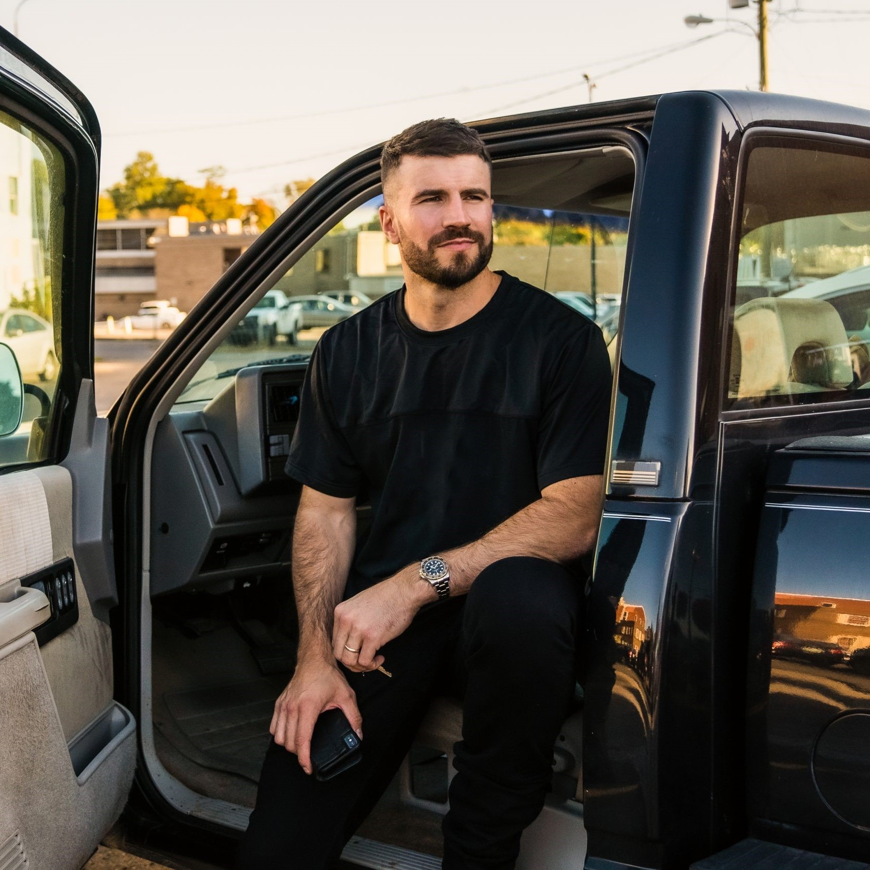 Sam Hunt, a bearded man with dark, cropped hair, sits in the open doorframe of a black pickup truck.