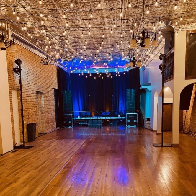 A medium sized room with walls of tan brick. Against one wall, a dark stage is set up, illumunated with bright blue and warm yellow string lights.
