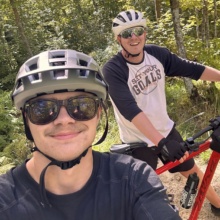 WCT writer Paul and a friend wear sunglasses and a helmet as they smile and bike through a heavily wooded area.