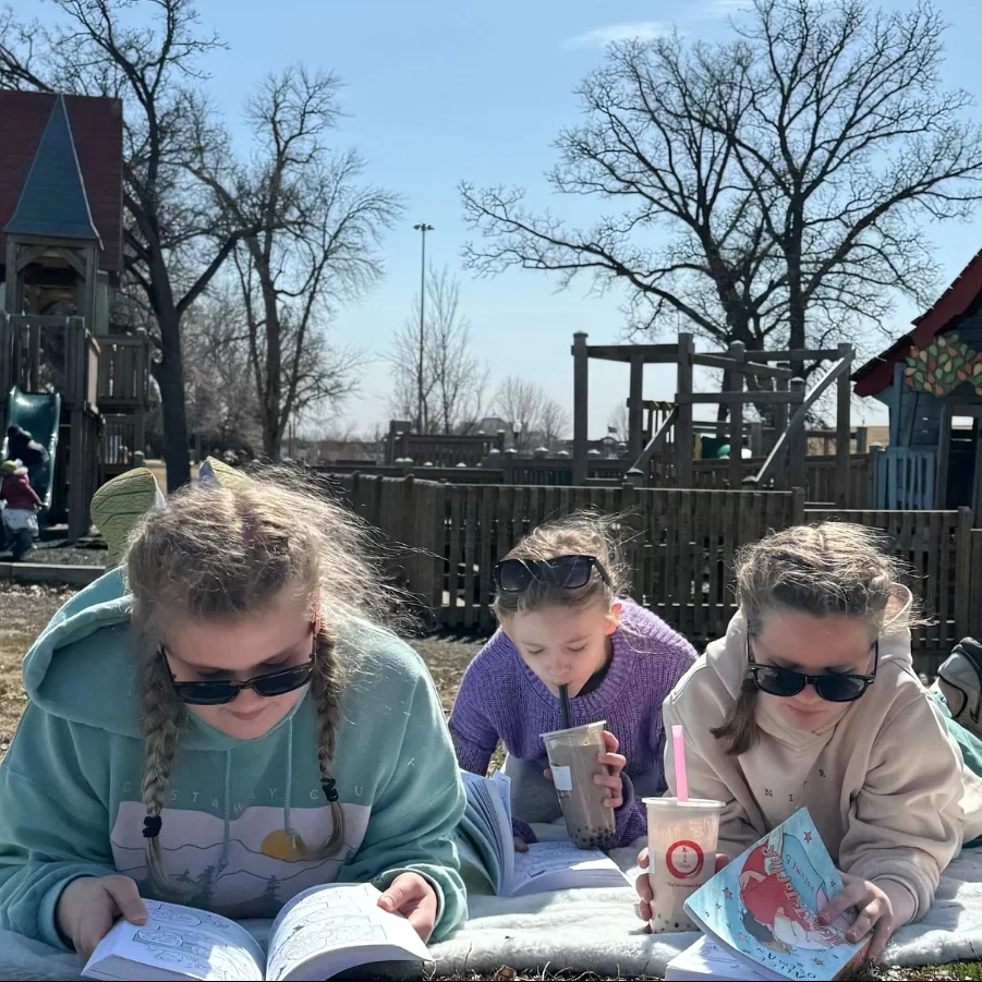 Three girls lay with Tea and Crepe beverages reading on a blanket in a Grand Forks park.