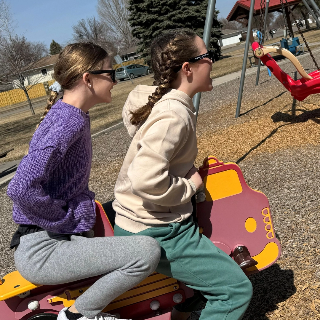 Two girls sit on a spring rider at the park with swings in view.