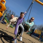 Three school-aged girls smile to the camera as they share a tire swing at a colorful park.