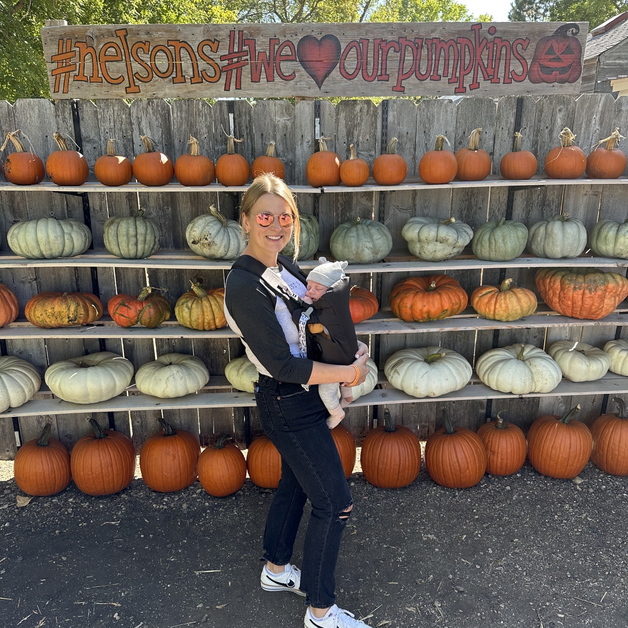 Author Jasmine holds her baby close to her chest in front of a wall of pumpkins at Nelson's Pumpkin Patch. Above them is a hand-painted sign that reads "#nelsons #weloveourpumpkins"