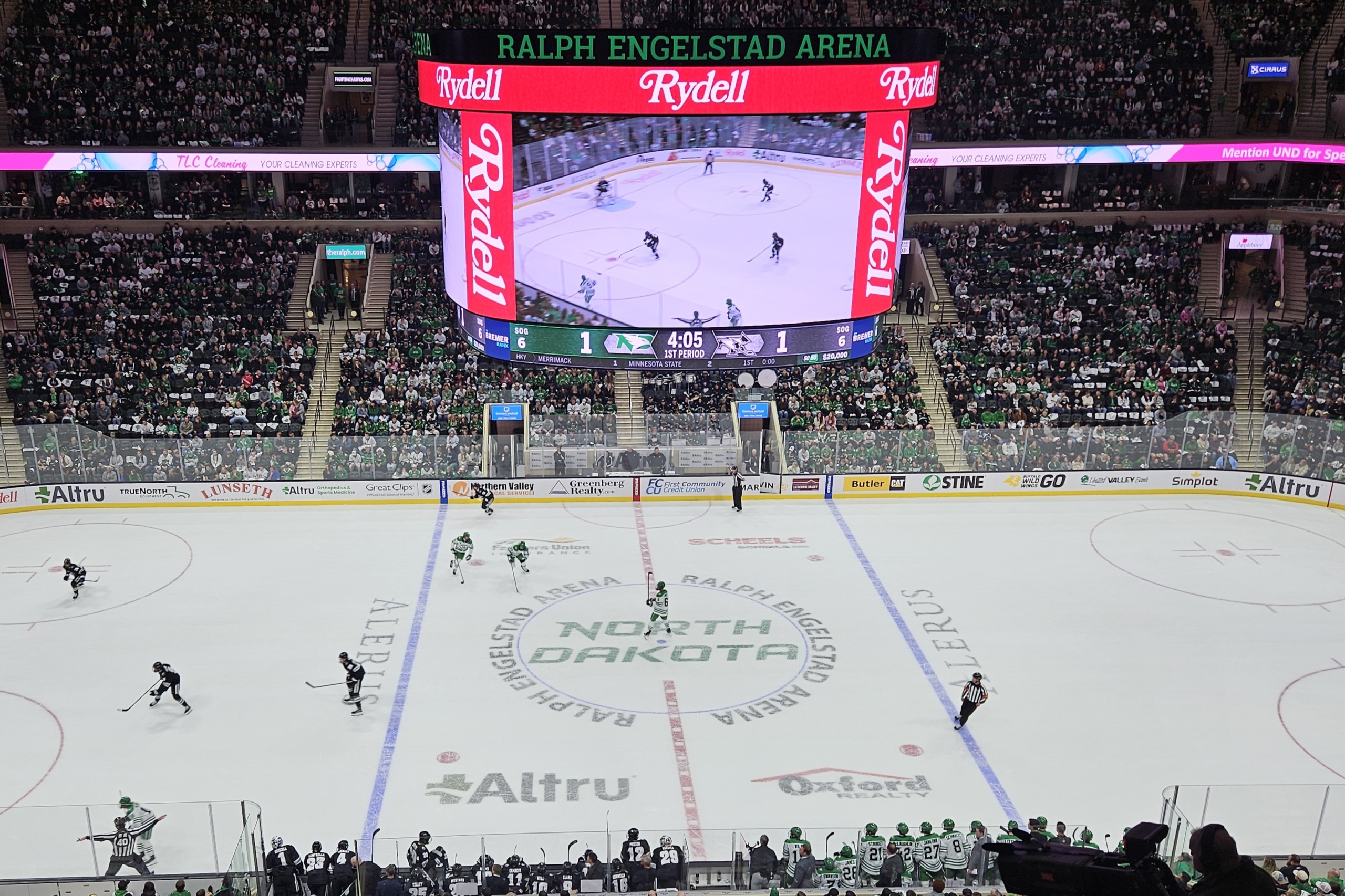 An aerial view of a UND hockey game in action, the massive scoreboard above displaying a score of 1 to 1. The seats of the Ralph Engelstad Arena are almost completely full.