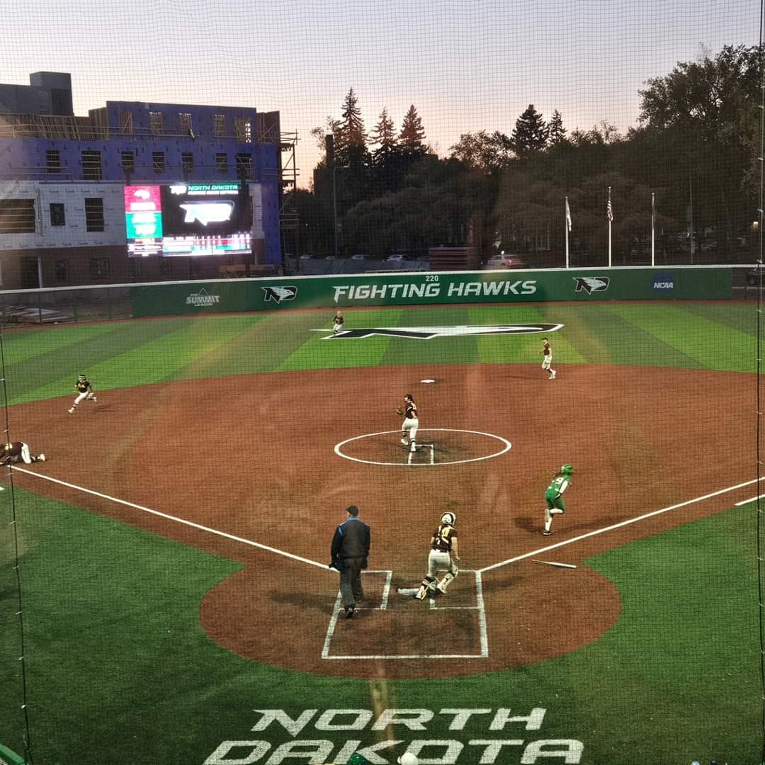 A softball game at Albrecht Field plays on at dusk. In the background is the large scoreboard as well as the framing of the new apartment complex being built near the field.