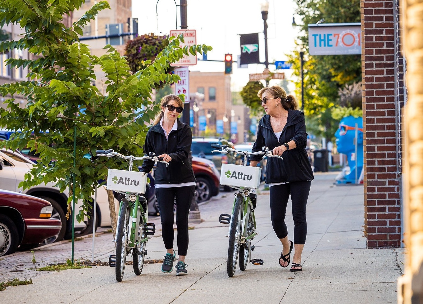 Two women walking downtown with the altru foundation city bikes. They are looking toward each other and laughing.