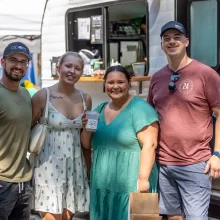 Picture of four adults at the downtown market in the summertime. Two men on the outsides and two women on the insides. Smiling at the camera in front of a little camper.