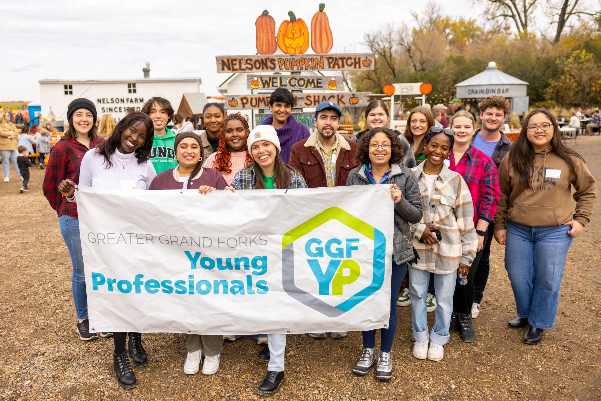 Author Gon Carlson stands in the middle front of a large group of young professionals at Nelson's Pumpkin Patch. They hold the GGFYP logo banner.