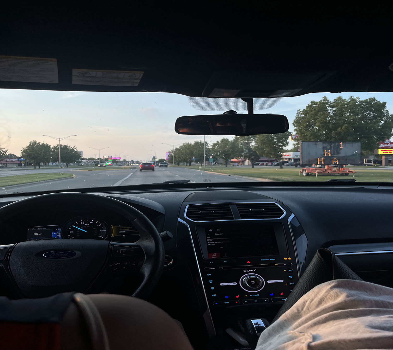 The camera sits in the front of a vehicle viewing outward from the windshield. Our view is of seemingly a summery evening as dusk is settling in. There is a large construction sign to the right of the image.