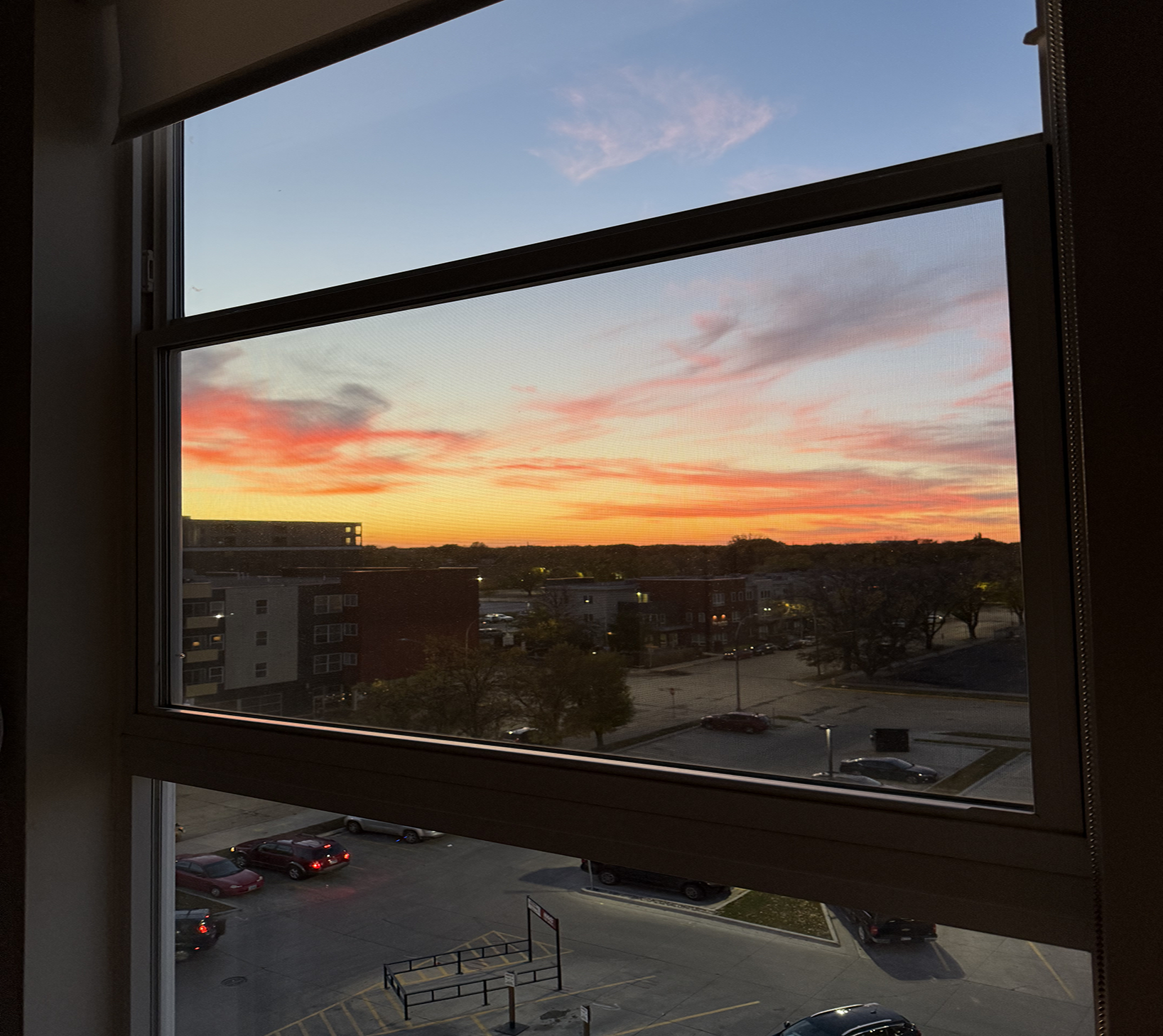 This image is through the window of an apartment building. It looks out onto a sunset from Grand Forks. The colors of orange, yellow, and pink bleed into the evening sky.