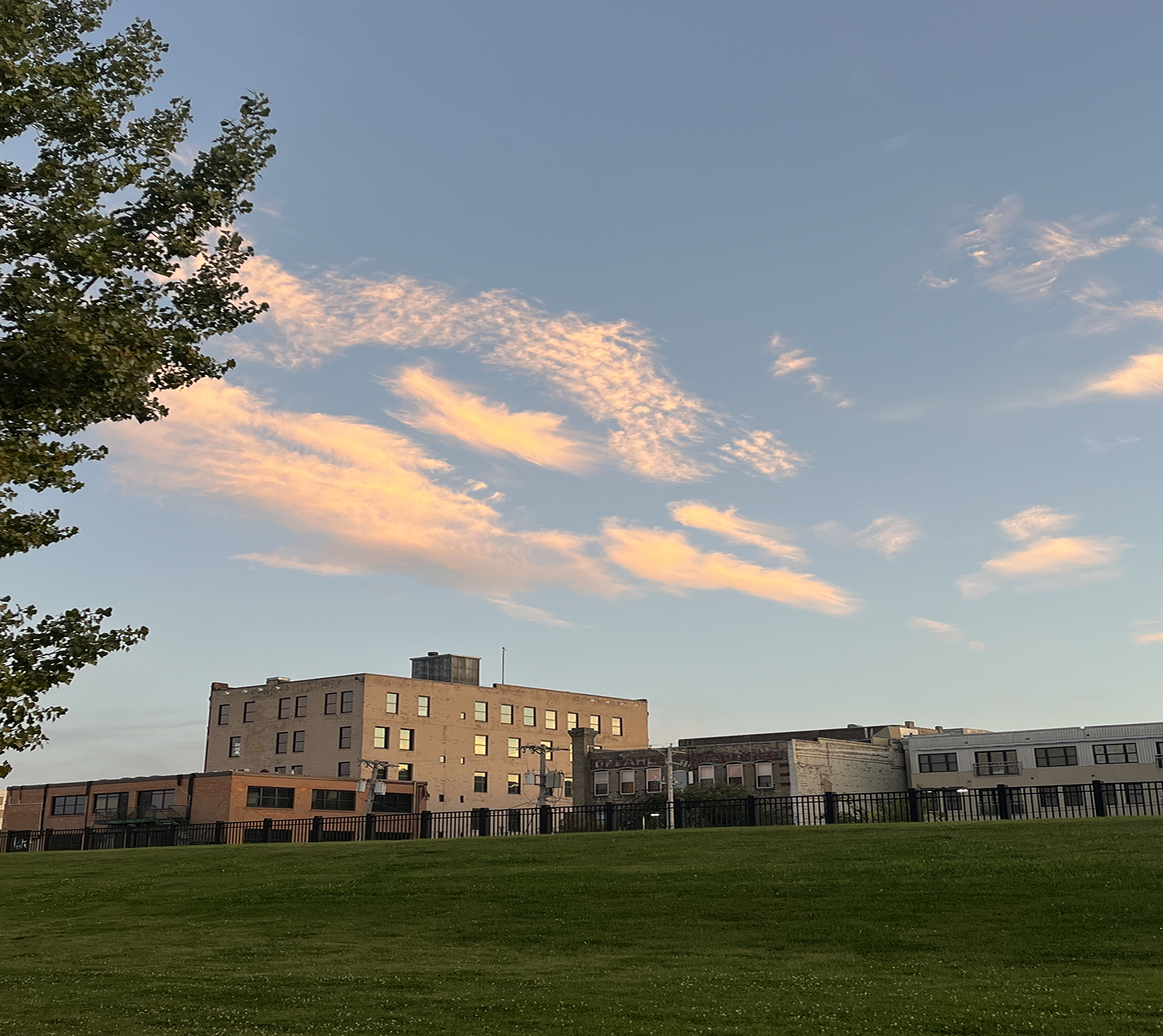 This image looks at a green field with a building in the back of the image. The dusk sky takes up the majority of the shot with colorfully, orange and pink lit clouds floating through a blue sky above.