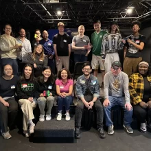 A large group of young professionals sit for a photo after their Mario Cart League event. They are in an indoor venue.