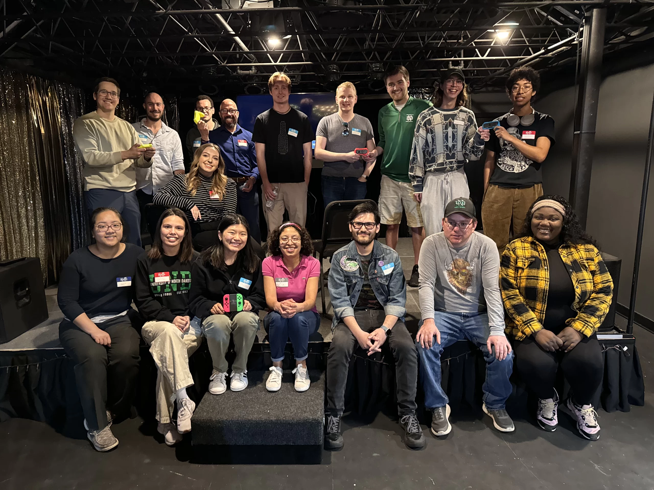 A large group of young professionals sit for a photo after their Mario Cart League event. They are in an indoor venue.