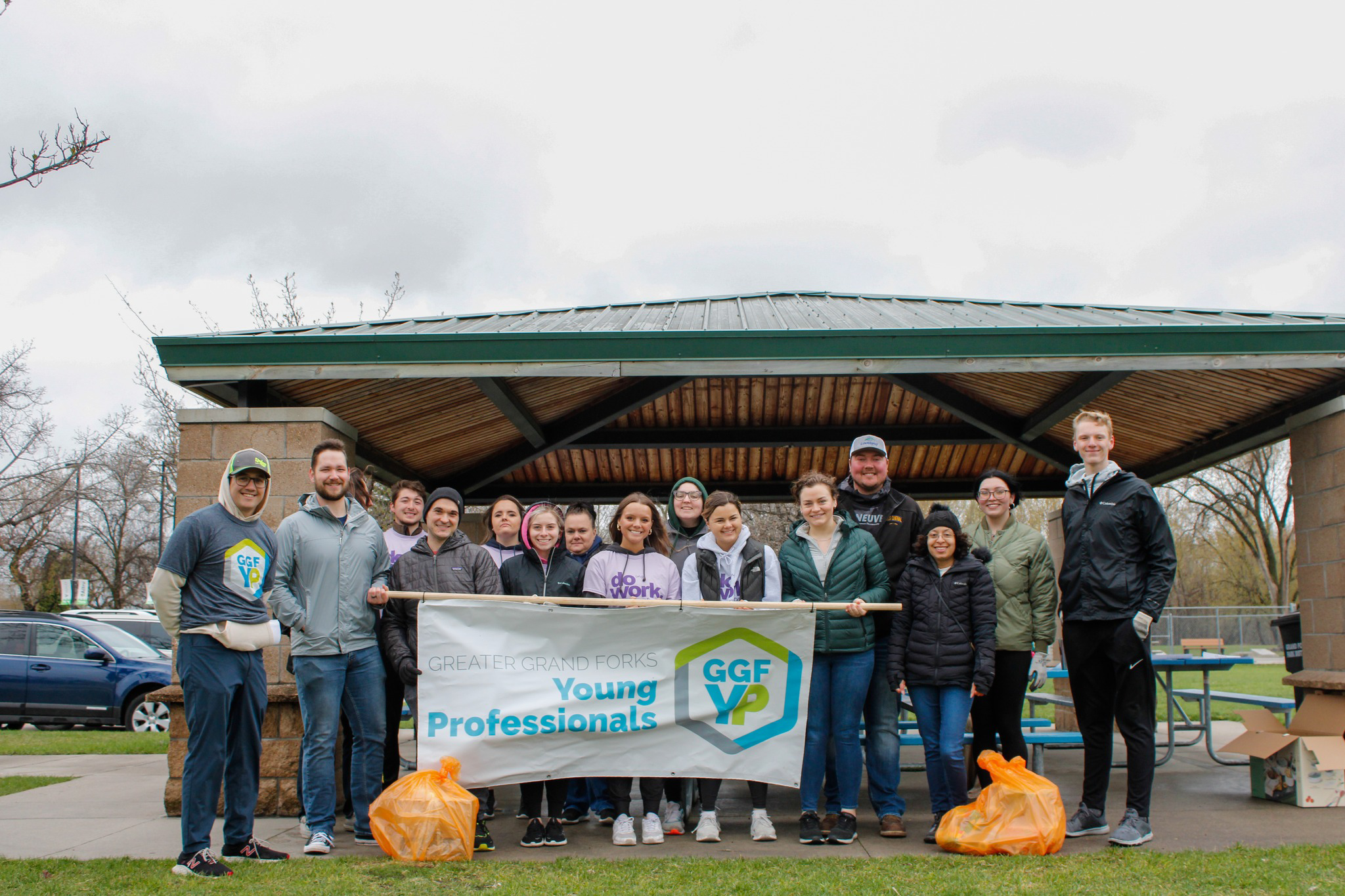 Young Professionals pose in a group with the GGFYP Banner for the Orange Bag Donations event. They are posing in front of an outdoor shelter in a park in Grand Forks on a dreary day.