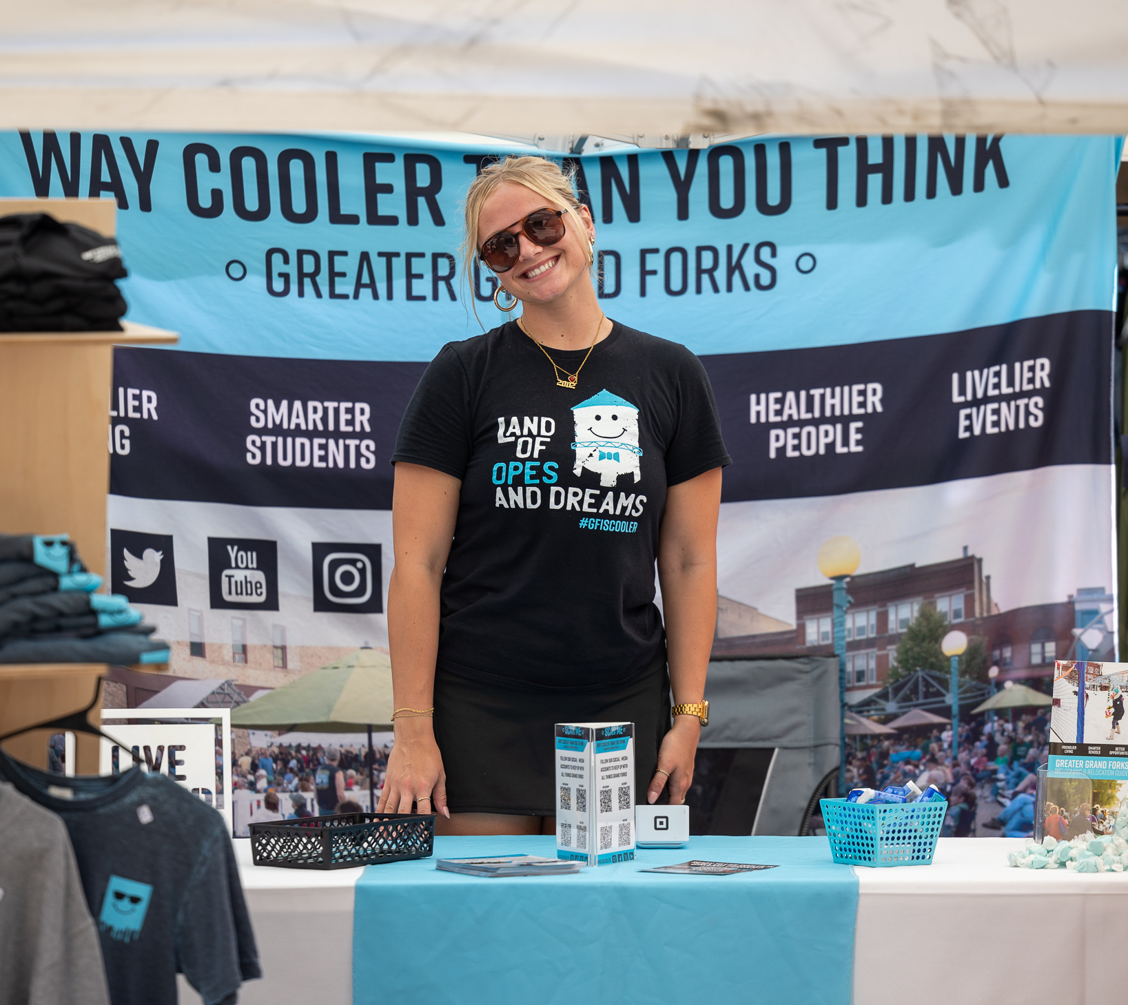 Author Isabelle stands at a Grand Forks is Cooler Merch booth at a summer market in Grand Forks. She stands smiling at the camera wearing a black "Land of Opes and Dreams" t-shirt.