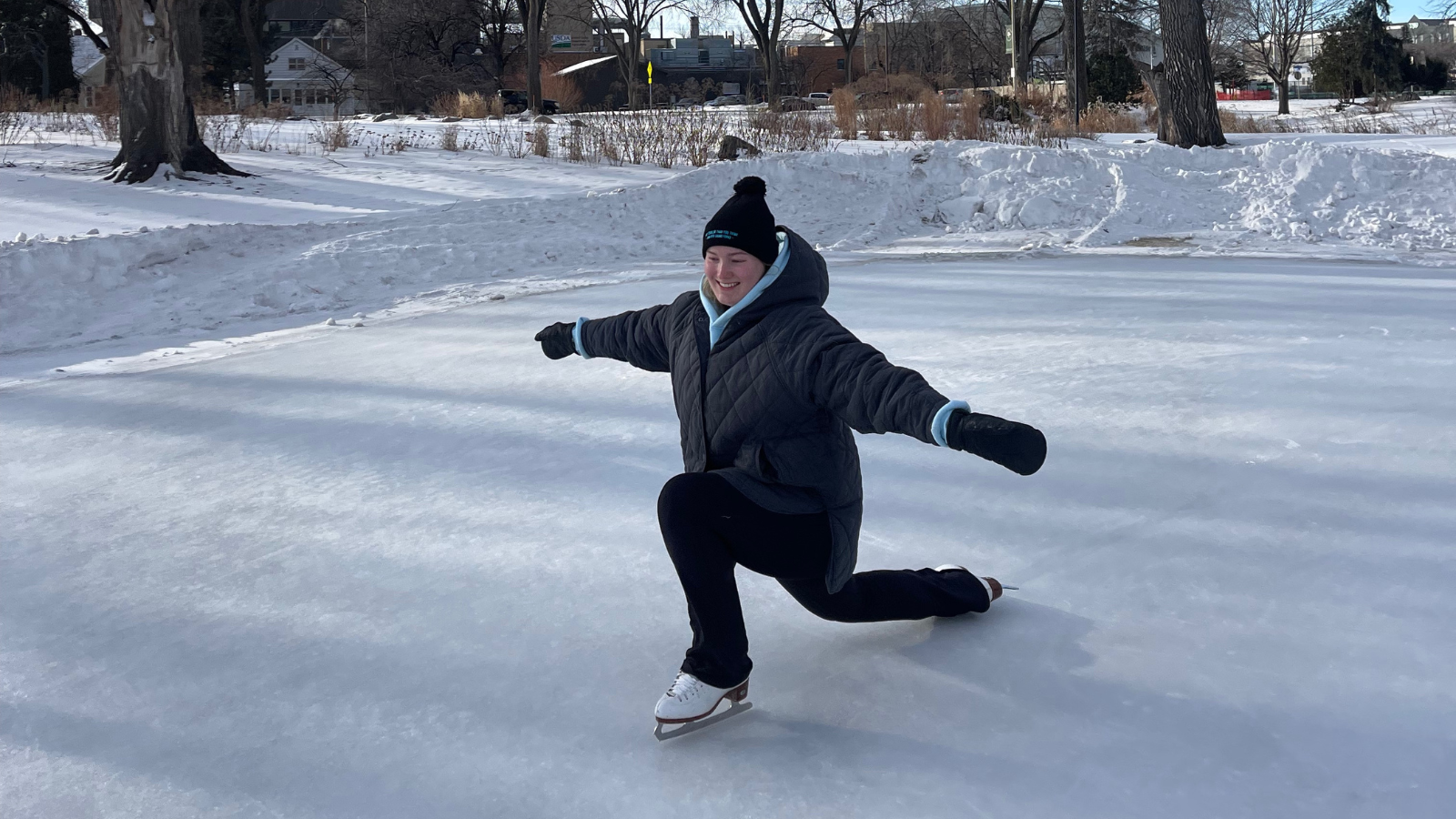 Author Kinley lunges down on one knee to show off her skating moves. She wears a big puffy grey coat and a big smile too.