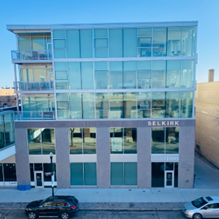 Glass walls surround the building on the top three floors. Glass balconies on the left hand side. Building is shorter with only five stories.