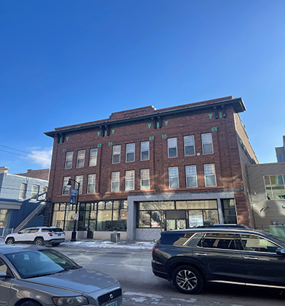 Picture of the historical building that is Stratford block off DeMers downtown. Red brick covering the facade with loads of windows and some green tiles with the letter S on them.