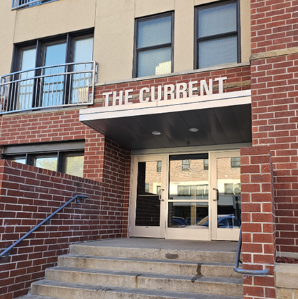 Front Entry Exterior of The Current Apartments. Lots of red brick and concrete steps. A big metal sign that reads "The Current" above the door hang.