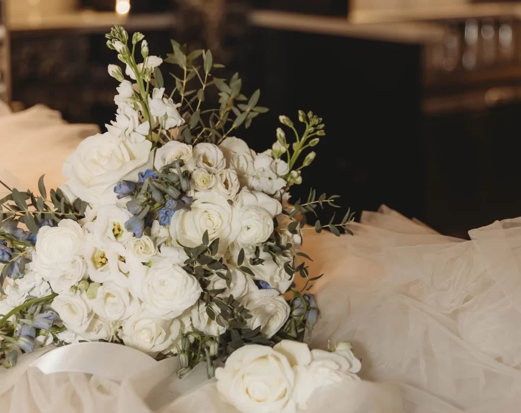 Pictured is a big white bouquet with white roses and hints of blue flowers as well. It sits on a cloud of white taffeta