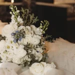 Pictured is a big white bouquet with white roses and hints of blue flowers as well. It sits on a cloud of white taffeta