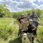 Family stands on the trails at Turtle River smiling at the camera. There are seven of them, including three adults at the back and four children in front with their yellow lab smiling big at the camera too.
