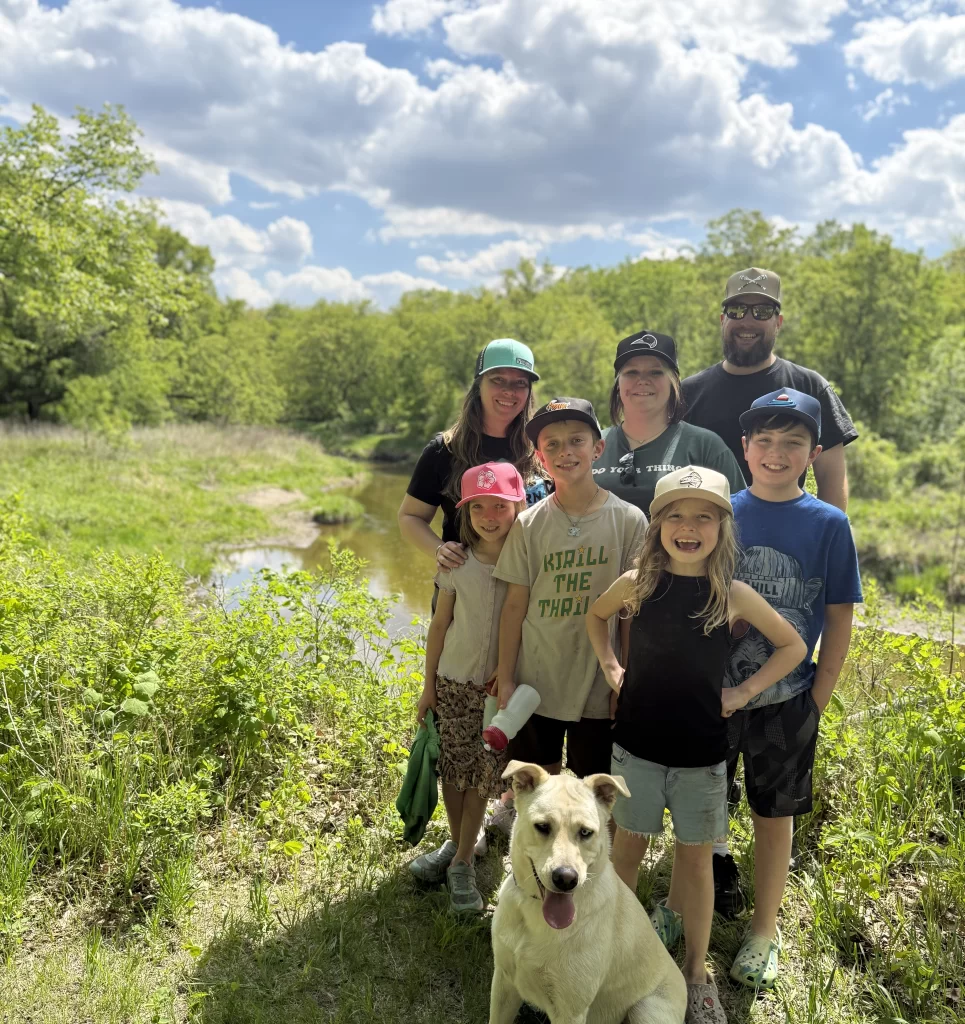 Family stands on the trails at Turtle River smiling at the camera. There are seven of them, including three adults at the back and four children in front with their yellow lab smiling big at the camera too.