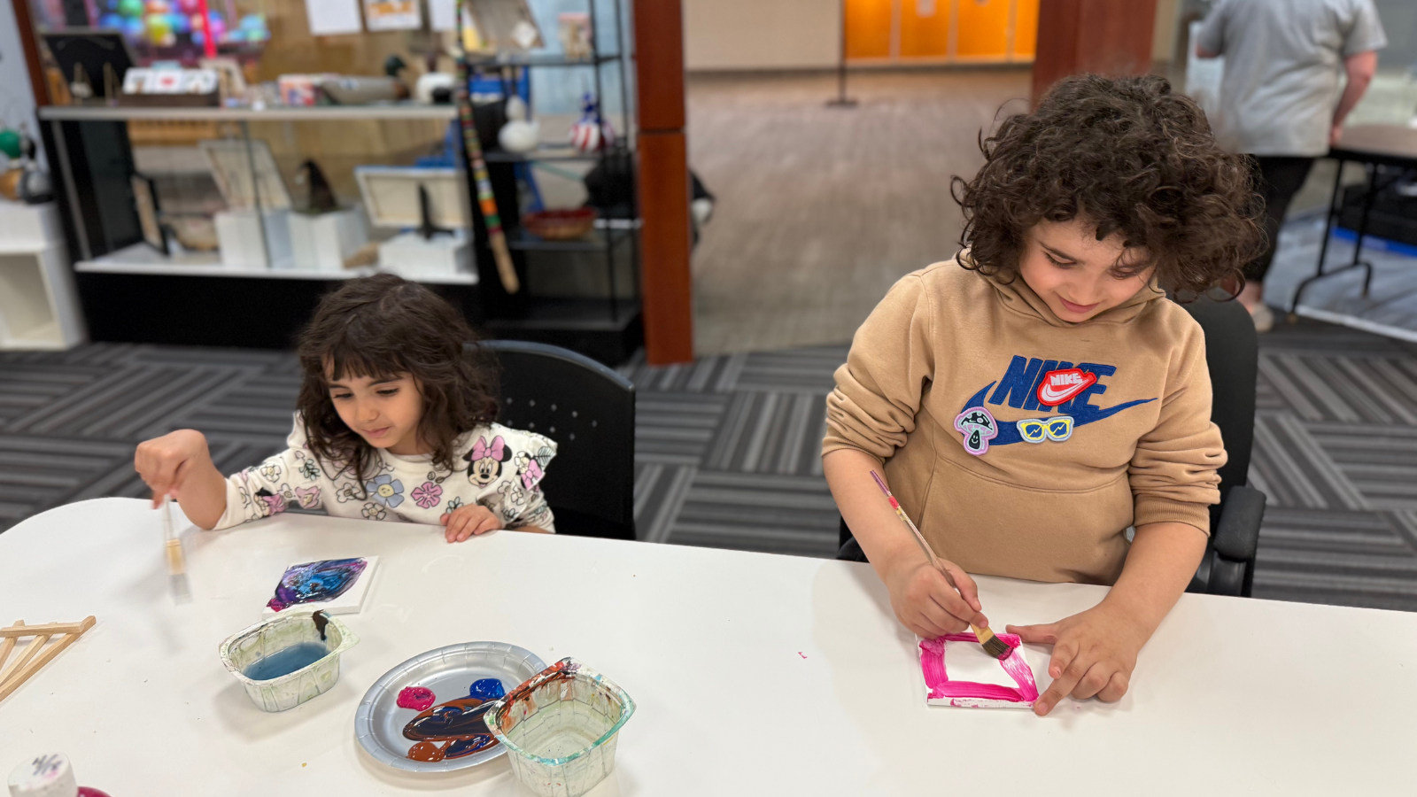 Kiddos work on their artful creations in this photo. They sit at a crafting table with all their supplies laid out in front of them.