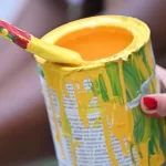 A pot of bright yellow paint is being dipped into with a paint brush. It is a very well loved pot of paint!