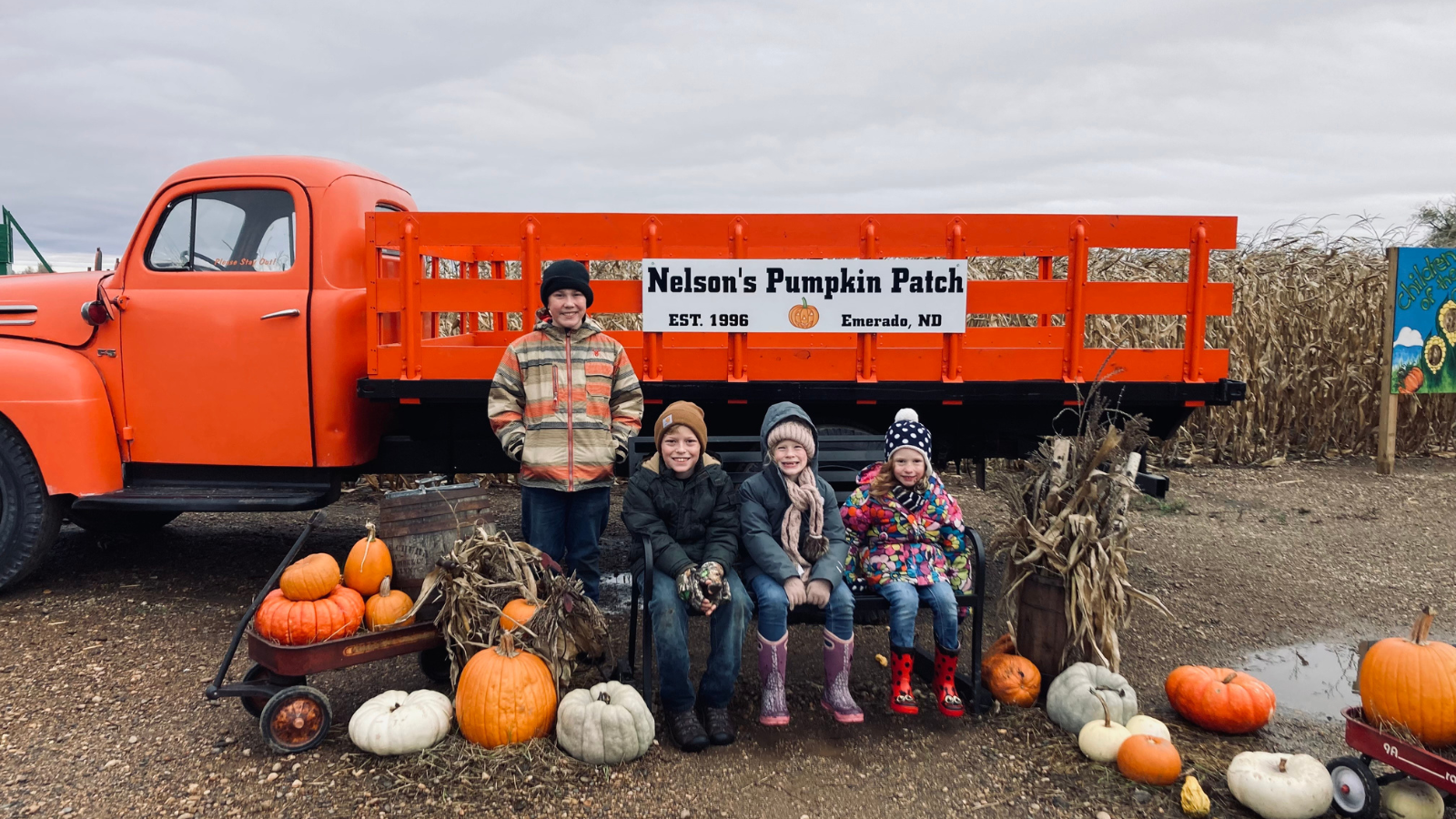 Nelson's Pumpkin Patch photo with four kiddos smiling at the camera sitting on a prearranged photo set with hay bales and pumpkins with a bright orange truck behind them.
