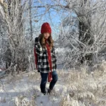 Author Natalie wears a black and white flannel with a matching red hat and scarf, as she stands in a scenic frosty winder landscape.