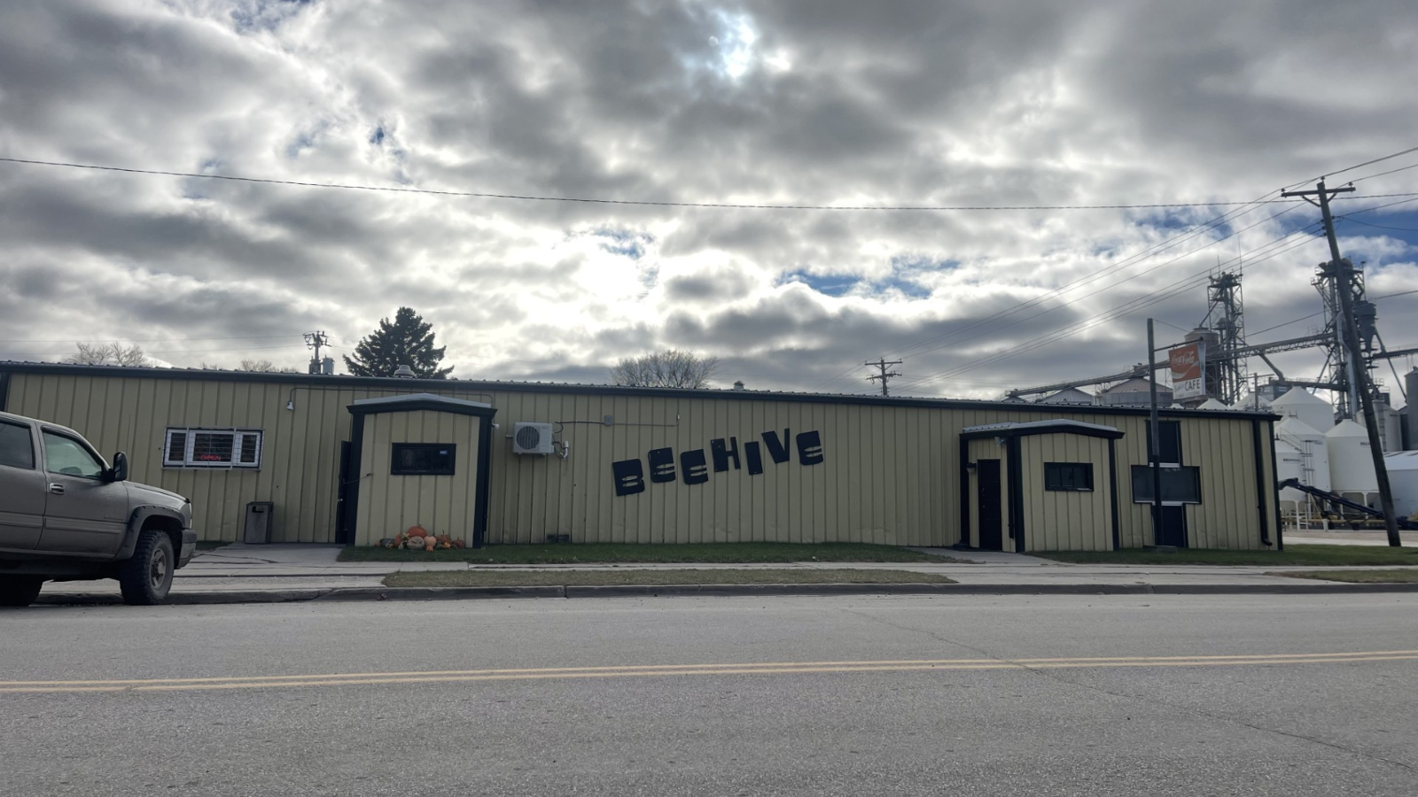Outside of the Beehive Bar in Reynolds, ND. The building is tan with brown trim on the outside and a the word "Beehive" spelled on the side of the building. The clouds have the sun shining through on the top half of the photo