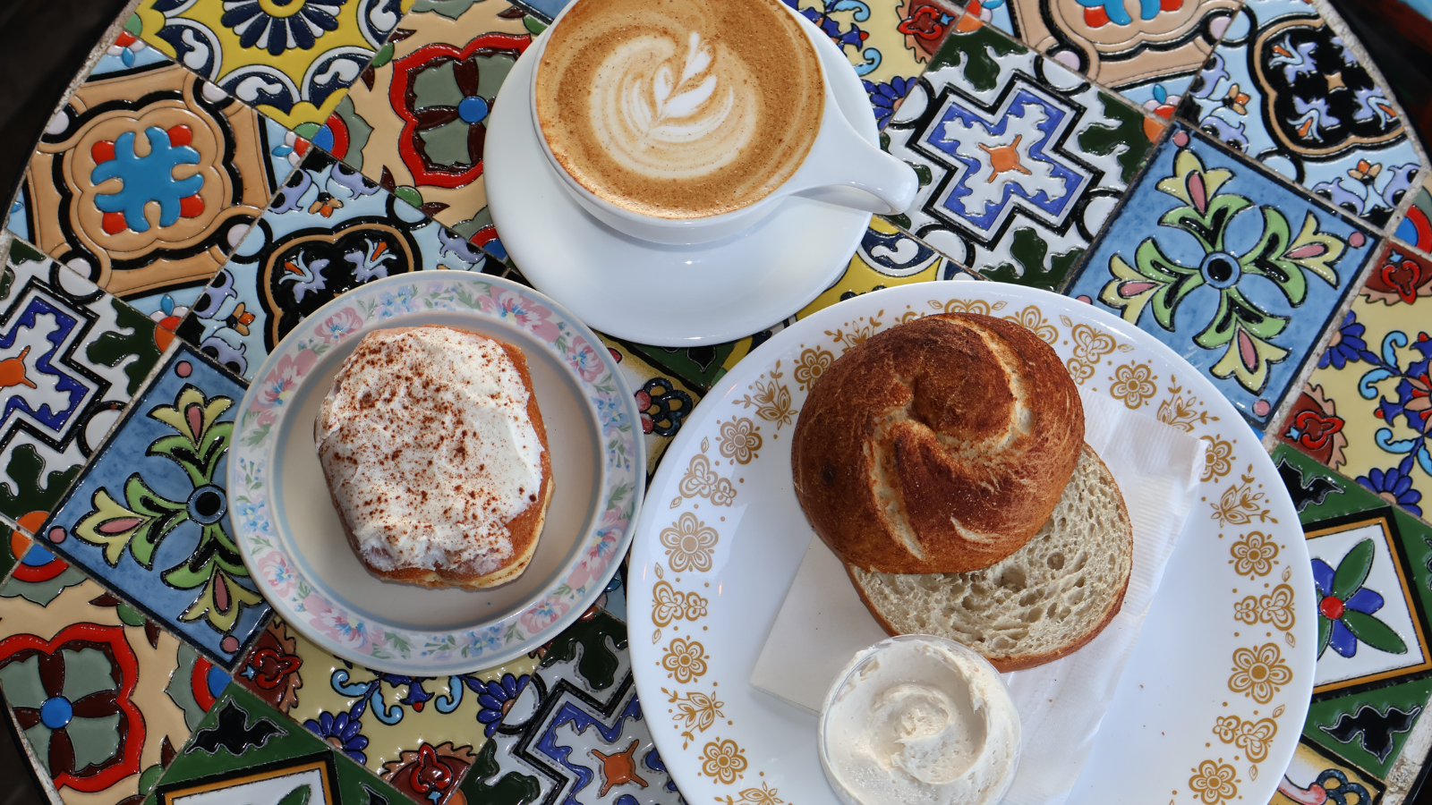An overhead image from Urban Stampede. Showcasing a latte, bagel, and cinnamon roll on a mosaic tiled table. yum!