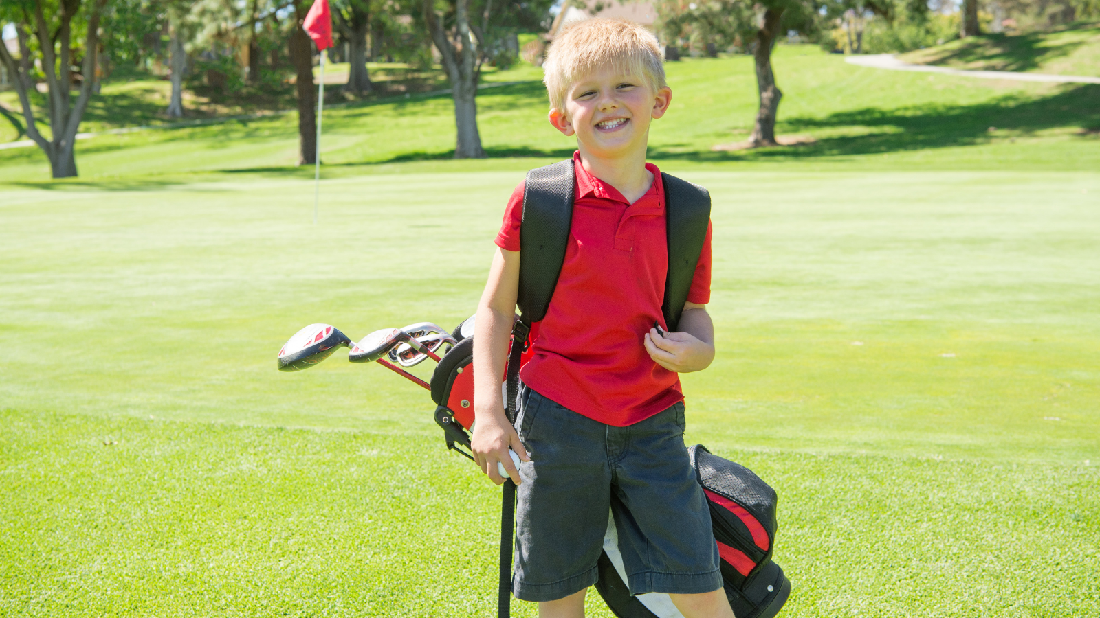 Golf Camp Kiddo Smiling at Camera Young boy smiles at the camera while holding golf clubs on the course