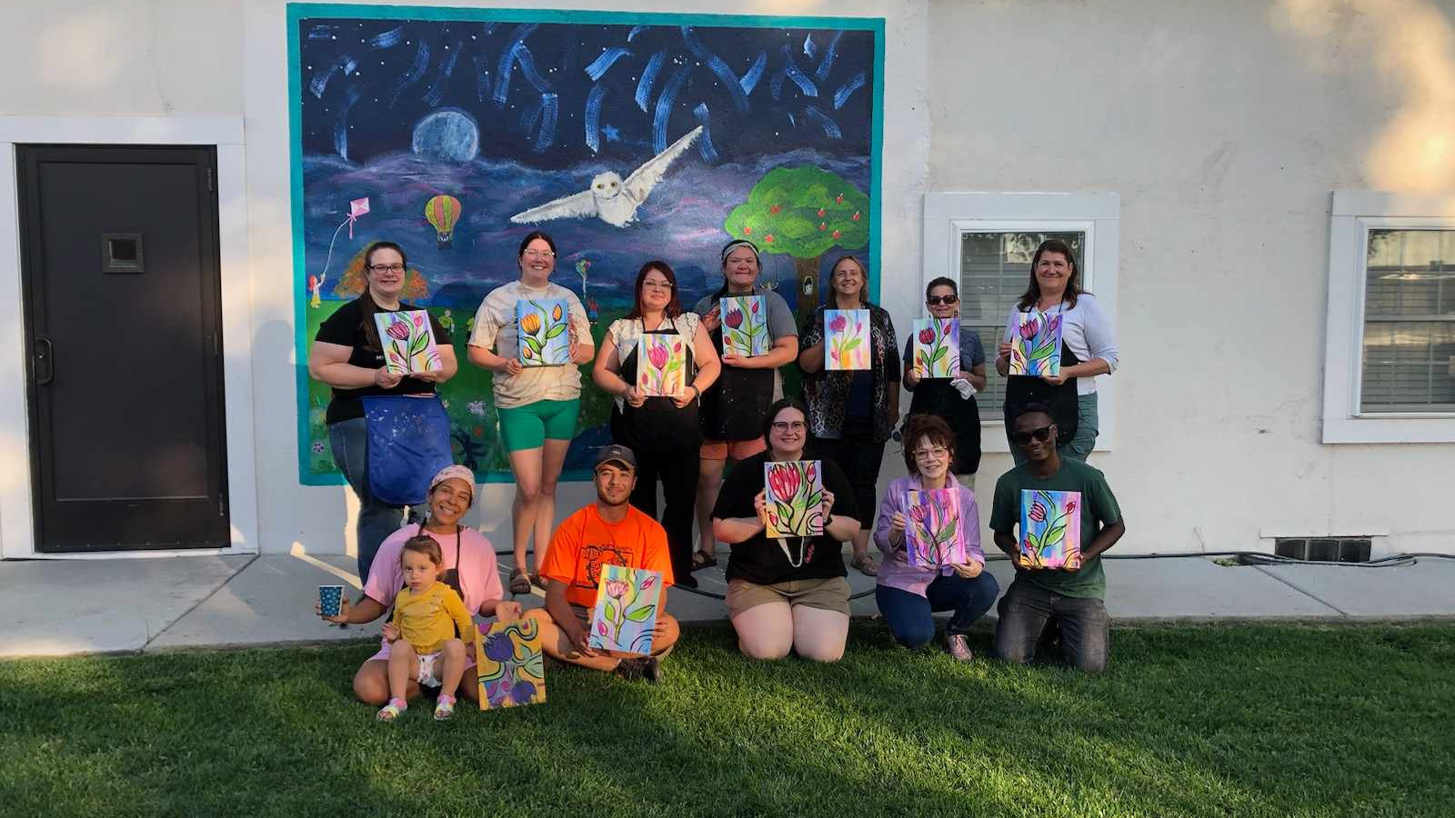 Artist Group Get Together Photo A multi-generational group stands in front of a mural with their individual art pieces in the summer in Grand Forks